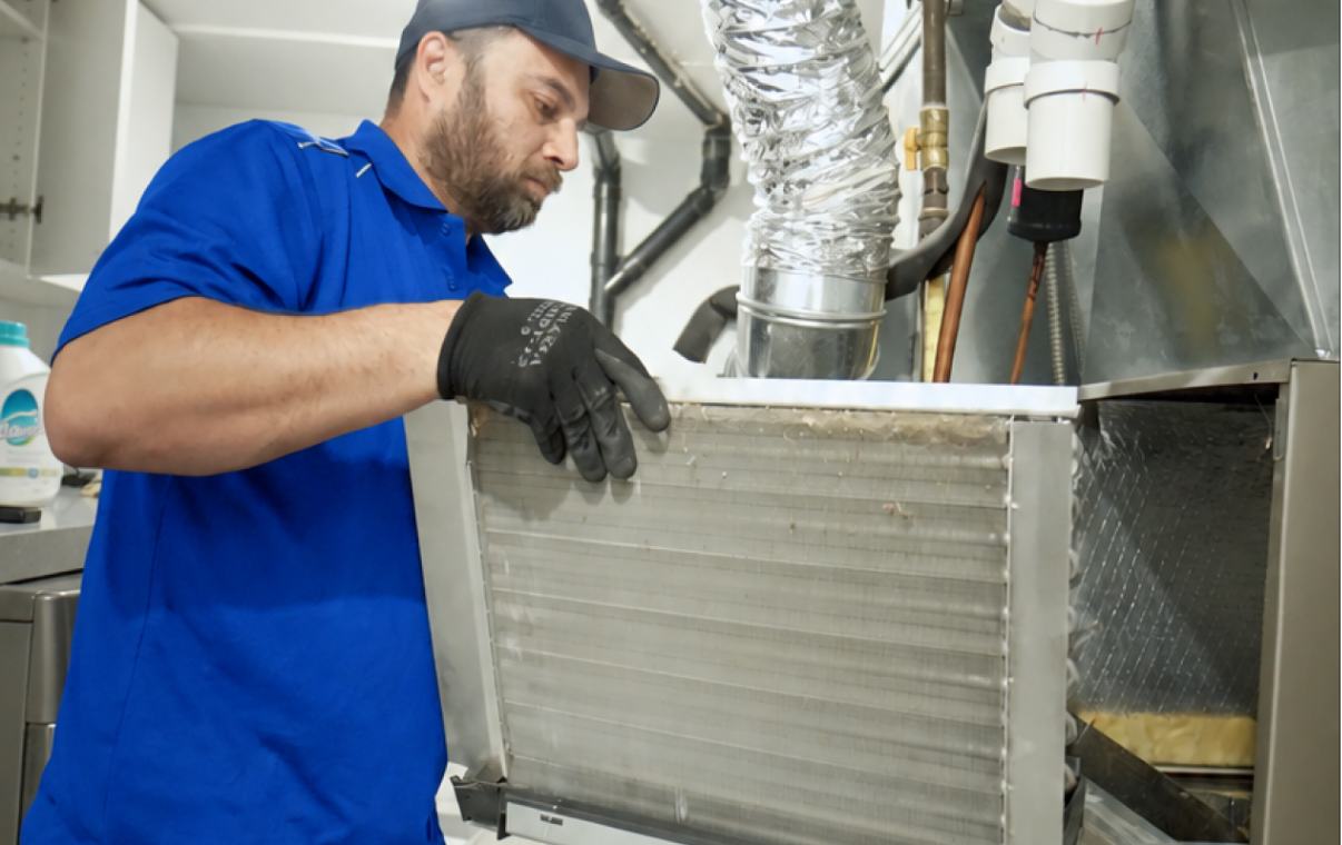 HVAC technician pulling a dusty evaporator coil from the cabinet above a furnace for inspection